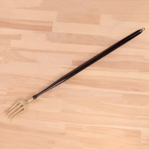 May include: A long, black handled brass fork with a ring at the end of the handle. The fork is lying on a light wood surface.