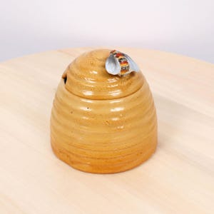May include: A golden yellow ceramic honey pot shaped like a beehive with a removable lid. A small, detailed ceramic bee with white wings and a striped body is attached to the top of the lid, serving as a handle for the honey container.