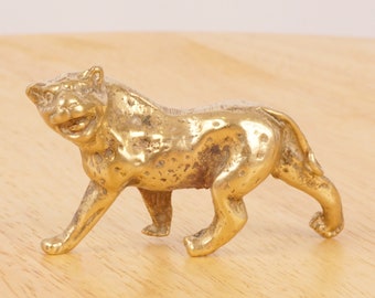 May include: A gold-colored brass figurine of a cheetah walking with its mouth open in a playful expression. The figurine is detailed with spots and has a textured surface.