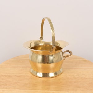 May include: A small, gold-colored brass bucket with a handle and a decorative design. The bucket is sitting on a wooden surface.
