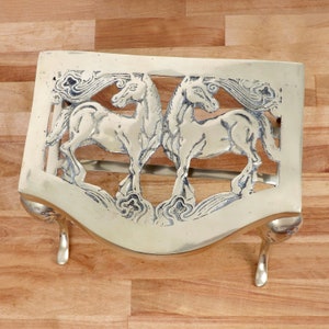 May include: A brass fire grate with two horses in a decorative design. The grate has three legs and a curved top.