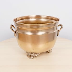 Puede incluir: Un cuenco de metal dorado en forma de caldero, con dos asas ornamentadas y tres patas decorativas. El cuenco tiene una parte superior ancha y abierta y un cuerpo redondeado. El metal tiene un acabado pulido. Para fines decorativos.