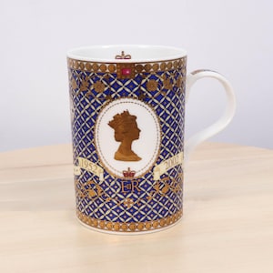 Może przedstawiać: Biały ceramiczny kubek z niebiesko-złotym wzorem. Kubek przedstawia sylwetkę osoby w koronie, z latami 1952 i 2002 po obu stronach. Kubek ma biały uchwyt i złoty brzeg.