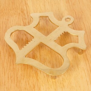 May include: A gold-colored metal trivet shaped like an anchor. The trivet has a decorative design with a serrated edge.