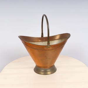 May include: A copper and brass coal scuttle with a handle. The scuttle has a decorative beaded rim and a brass base.
