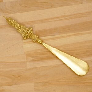 Peut inclure: Un chausse-pied en m&eacute;tal dor&eacute; avec une poign&eacute;e d&eacute;corative et orn&eacute;e en forme de tour. Le chausse-pied est sur une surface en bois brun clair.