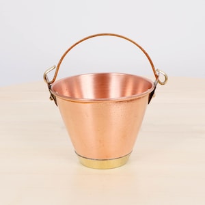 May include: A small, copper-colored bucket with a brass base and a leather handle. The bucket has a rounded shape and is designed with a sturdy handle for easy carrying. The metal has a polished finish, reflecting light.