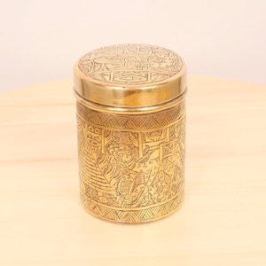 May include: A gold cylindrical container with an intricate engraved design featuring a pyramid and animals. The lid is also engraved with a similar design.