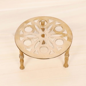 May include: A gold-coloured metal trivet with a circular design. The trivet has a decorative openwork pattern and three short legs. The trivet is designed to protect surfaces from hot cookware.