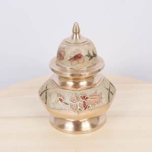 May include: A gold-colored decorative jar with a lid. The jar features a floral design in shades of red, brown, and cream. The jar is sitting on a light-colored wooden surface.