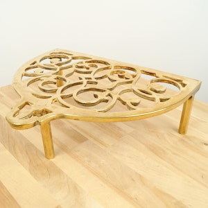 May include: A gold-coloured metal trivet with a decorative, intricate design. The trivet has three legs and is shaped like a half-circle.