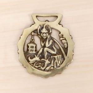 Könnte beinhalten: Ein Messing-Pferdegeschirr mit einem detaillierten Relief eines sitzenden Dämons. Der Dämon, mit Hörnern und spitzem Kinn, ist von einem dekorativen Rand und einer hängenden Laterne umgeben. Das Messing hat eine warme, goldene Farbe.