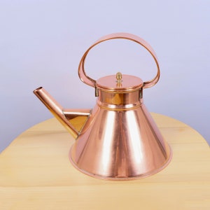 Op de afbeelding: Een koperen theepot met een glanzende afwerking en een rond, kegelvormig lichaam. De theepot heeft een enkele handgreep en een deksel met een kleine knop.