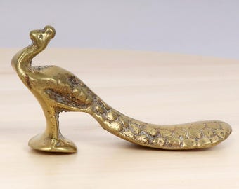 Puede incluir: Una figurita de pavo real de latón dorado con una cola texturizada y una cabeza levantada. La figurita está de pie sobre una superficie de madera.