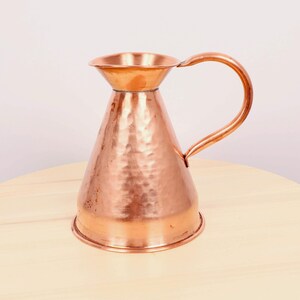 May include: A hammered copper jug with a wide mouth and a curved handle. The jug has a shiny, metallic finish.