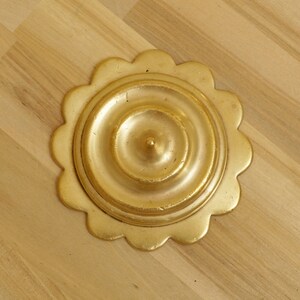 Könnte beinhalten: Eine goldfarbene, dekorative Rosette mit einem gewellten Rand. Die Rosette hat ein konzentrisches Kreisdesign mit einer erhabenen Mitte.