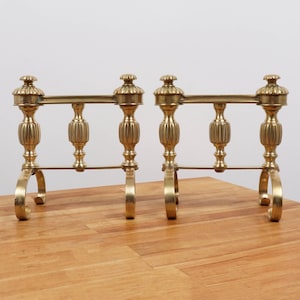 May include: A pair of matching brass fireplace andirons featuring a horizontal bar supported by three vertical, fluted, spindle-shaped columns. Each andiron is topped with a decorative finial and rests on two curved, arched metal feet.