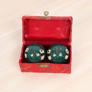 May include: A pair of green Chinese health balls with panda and bamboo designs, presented in a red, fabric-lined box. The box is rectangular with a gold-colored clasp. The balls are approximately 2 inches in diameter.