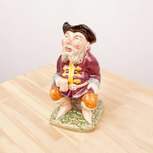 Puede incluir: Una figurita de cerámica de un hombre que lleva una chaqueta roja y dorada, un sombrero negro y sostiene una taza. Está sentado sobre una base verde con textura de piedra.