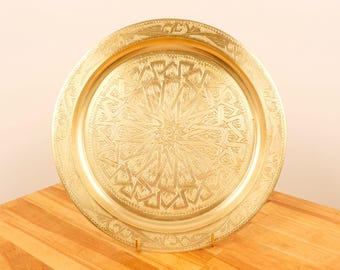 May include: A round, gold-colored metal serving tray with an intricate, embossed design. The tray has a raised rim and a decorative border.