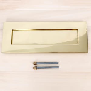 May include: A rectangular, polished brass letterbox with a recessed mail slot. Two silver-colored bolts with brass-colored heads are positioned below the letterbox. The letterbox is a bright, shiny gold color.