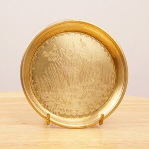 Könnte beinhalten: Eine goldfarbene Metallschale mit einem geätzten Design eines Kobolds, der einen Krug hält. Die Schale hat einen gewellten Rand und die Worte "Lucky Pixie" sind um den Boden herum eingraviert.