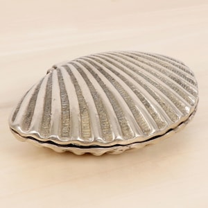 May include: A silver-coloured, clam-shaped trinket box with a hinged lid. The box has a detailed, ridged texture, resembling a real seashell. The box is closed and sits on a light-coloured surface.