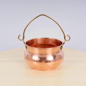 May include: A small copper pot with a hammered finish and a brass handle. The pot is sitting on a wooden surface.