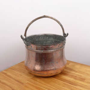 May include: A copper bucket with a brass handle. The bucket has a rounded bottom and a slightly flared top. The bucket is old and has a patina of age.