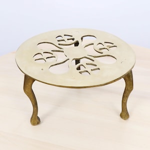 Puede incluir: Un salvamanteles redondo de color latón con un diseño decorativo recortado. El salvamanteles tiene tres patas curvas y una parte superior circular con un patrón floral. Está diseñado para proteger las superficies de los objetos calientes.