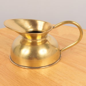 Könnte beinhalten: Ein polierter Messingkrug mit einem breiten, ausgestellten Ausguss und einem abgerundeten Körper. Der Krug hat einen einzigen Henkel und eine glatte, glänzende Oberfläche.