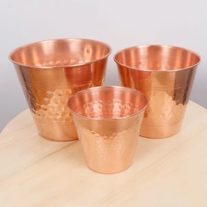 May include: Three copper-coloured metal planters of different sizes. Each planter has a hammered texture and a flared rim. The planters are arranged on a light-coloured wooden surface, highlighting their metallic sheen.