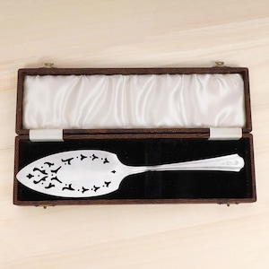 Puede incluir: Un servidor de pastel plateado con un diseño calado decorativo, presentado en un estuche marrón vintage con un interior de terciopelo negro y forro de satén blanco. El servidor tiene un mango largo y una hoja detallada.