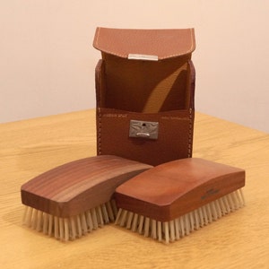 Könnte beinhalten: Zwei Holzschuhbürsten mit Naturborsten und ein braunes Lederetui mit den Aufschriften "PIGSKIN SPLIT" und "MADE IN ENGLAND".