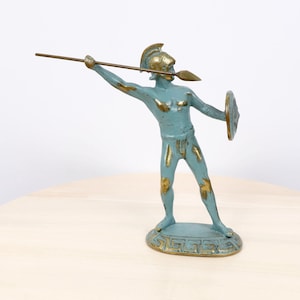 May include: A metal figurine of a warrior in a helmet, holding a long spear and a round shield. The figure has a blue-green finish with worn gold-toned highlights on the helmet, spear, shield, and base, which features a Greek key pattern.