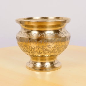 May include: A gold metal vase with an intricate floral design etched into the surface. The vase has a wide mouth and a rounded base.