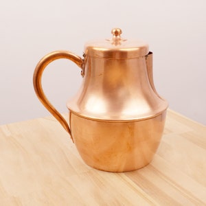 Puede incluir: Una jarra de cobre con cuerpo redondeado y tapa. La jarra tiene un asa y un acabado liso y brillante.