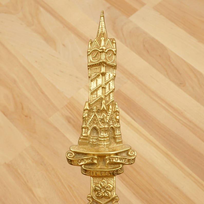 Peut inclure: Ornement en m&eacute;tal dor&eacute; en forme de tour avec un cadran d'horloge et les mots "Clock Tower Leicester" inscrits sur une banni&egrave;re.