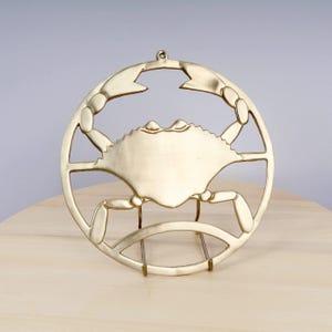 May include: A gold-coloured metal crab-shaped hot pad with a round design. The hot pad has a stand on the bottom.