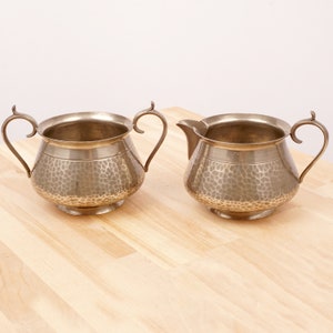 May include: A set of two silver-toned hammered metal creamer and sugar bowls. The bowls have a rounded shape with a hammered texture and a shiny finish.
