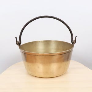 Puede incluir: Un caldero de latón vintage con un asa de metal oscuro. El caldero tiene un cuerpo redondeado y un borde ligeramente ensanchado. El asa es arqueada y está sujeta a los lados. El latón tiene un tono dorado cálido.