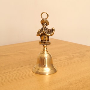 Pode incluir: Um sino de latão com uma figura de um homem de chapéu e casaco. A figura está acima do sino, com o texto "RIPON HORNBLOWER" numa pequena placa. O sino tem uma argola no topo para pendurar.