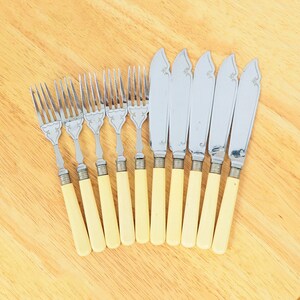 May include: A set of ten silver-plated butter knives with ivory-coloured handles. The knives are arranged in a row on a wooden surface.