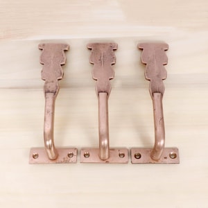 May include: Three copper-colored metal hooks with a decorative top and a curved handle. Each hook has a rectangular base with three screw holes. The hooks are arranged in a row on a light wood surface.