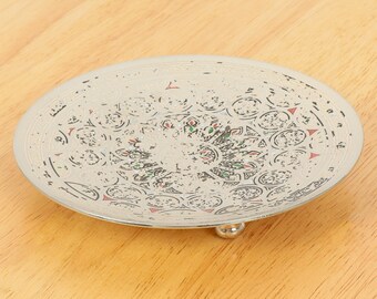 May include: A silver decorative tray with an intricate design featuring circles, triangles, and floral patterns in black, red, and green. The tray has three small feet.