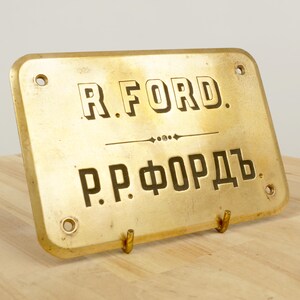May include: A brass plaque with the text "R. FORD." and "P.P. ФОРДЪ" in black lettering. The plaque has two holes on the top and two on the bottom, and is mounted on a wooden stand.