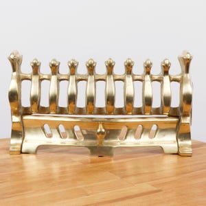 May include: A polished brass fireplace grate featuring a series of vertical bars topped with finials. The base is constructed with a solid horizontal plate containing decorative cutouts and a central knob, providing a sturdy structure.