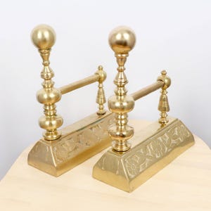 Puede incluir: Par de atriles de chimenea antiguos de latón. Cada atril presenta un remate en forma de bola, una columna decorativa y una barra horizontal. La base rectangular tiene relieves florales.