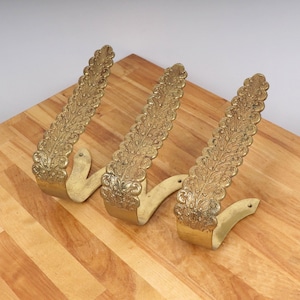 May include: Three ornate brass wall hooks with a leaf-like design. The hooks are a warm gold color and are mounted on a light-colored wooden surface. The hooks have a curved shape with a decorative, textured pattern.