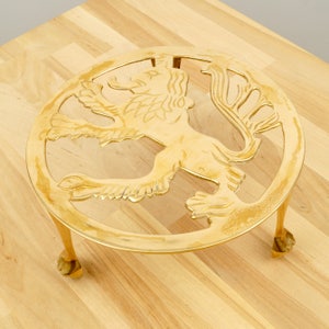May include: A gold-toned metal hot pad featuring a lion design. The hot pad has three legs and is round.